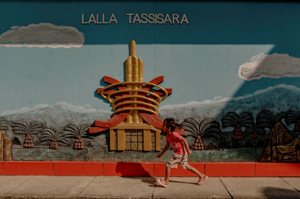 Seorang anak bermain didekat mural Lalla Tassiara yang dihiasi lukisan sawit. Lalla Tassiara berarti "bersatu atas keragaman" yang digunakan sebagai semboyan Kabupaten Mamuju Tengah. Foto: Iqbal Lubis/Bollo.id